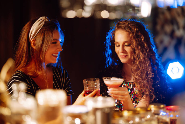Cheerful women drinking cocktails in bar. Women friends make a toast as they celebrate at a party. Party, celebration, friends, bachelorette party, birthday, winter holidays concept. - 写真・画像