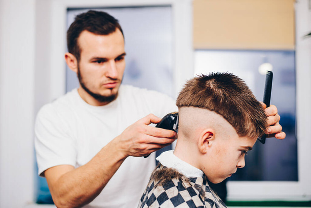 young barber making haircut of cute little boy at salon - Foto, afbeelding