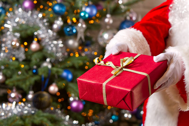 Santa Claus hands holds red gift box opposite christmas tree. - Фото, изображение