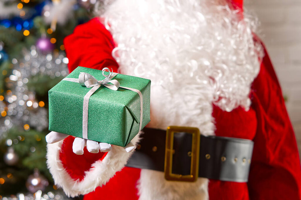 Noel Baba elleri Noel ağacının karşısındaki yeşil hediye kutusunu tutuyor.. - Fotoğraf, Görsel