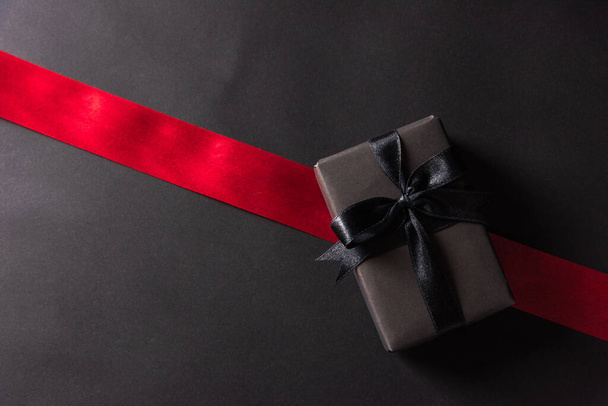 Black Friday Sale concept, Top view of gift box wrapped black paper and black bow ribbon press, studio shot on and having red ribbon across a dark background - Foto, imagen