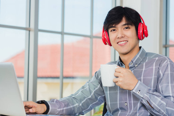 Asiatischer junger Geschäftsmann glücklich mit roten Kopfhörern am Schreibtisch sitzend, Home Office mit Laptop, selbstbewusster gutaussehender Mann mit Lifestyle-Lächeln, entspannt eine Kaffeetasse haltend, Musik hörend - Foto, Bild