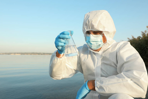 Scientist in chemical protective suit with conical flask taking sample from river for analysis - Foto, afbeelding