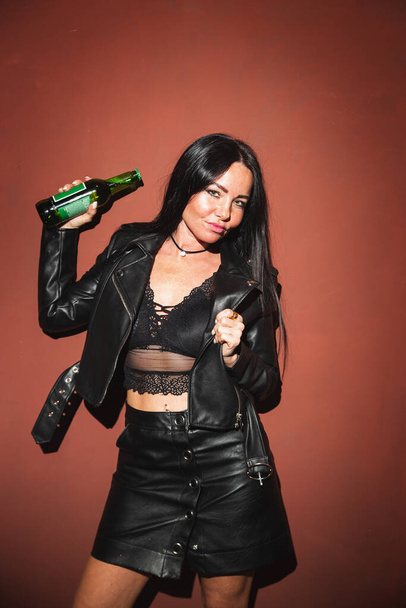 Sexy brunette woman in leather jacket leather skirt and lace bralette posing with beer red wall background hard light - Φωτογραφία, εικόνα