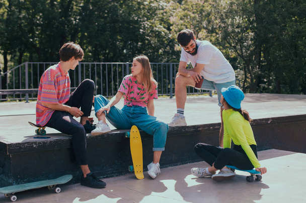 In een goede zonnige dag, groep vrienden zeer aantrekkelijke multi-ethnische genieten van de tijd in een moderne, skate park ze zitten op de vloer en socialiseren voordat u begint met skateboarden - Foto, afbeelding