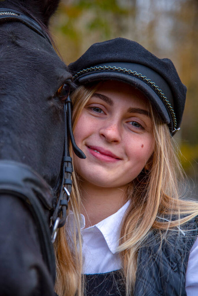 Portrait of a teenage girl with long blonde hair and a black cap, hugging a Chestnut horse against the background of an autumn forest. - Foto, imagen