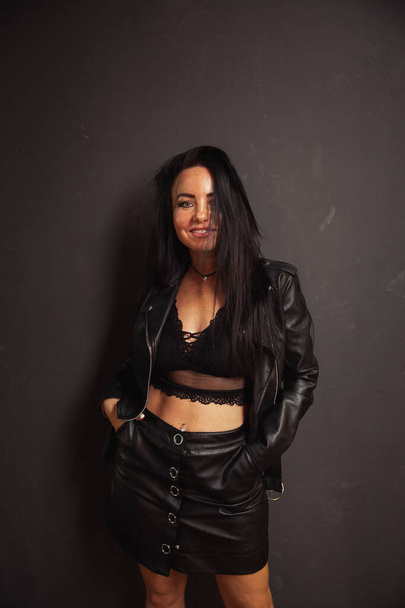 Sexy unformal brunette woman in leather jacket leather skirt and lace bralette posing dark wall background hard light - Foto, Imagem