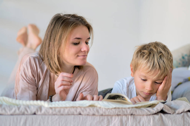 Η μαμά διδάσκει ότι ο γιος που διαβάζει ένα βιβλίο είναι στο κρεβάτι. Εκπαιδευτικές αντιλήψεις. Χρώματα παστέλ, επιλεκτική εστίαση. - Φωτογραφία, εικόνα