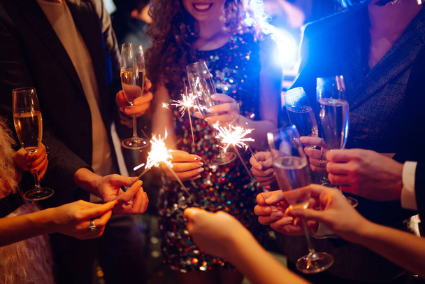 Sparkling sparklers in the hand. Group of happy people holding sparklers at the party. Young friends clinking glasses of champagne  in a nightclub. Celebration, people and holidays concept. - Photo, Image