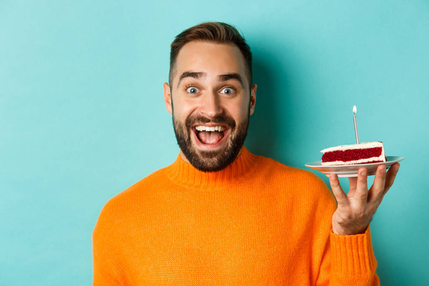 Detailní záběr šťastný dospělý muž slaví narozeniny, drží bday dort se svíčkou a přání, stojí na tyrkysovém pozadí - Fotografie, Obrázek