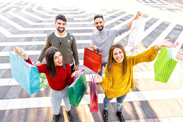 View from top of two couple outdoor in the city showing their colored shopping bags smiling and looking at the camera - Vivid colors of beautiful stylish young friends satisfied with purchases - Photo, Image