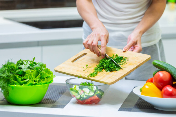 Close up of a girl hands cutting greenery with a knife on a cutting board for a vegan vitamin vegetable salad and putting them in a glass bowl while cooking breakfast in the kitchen - Photo, image