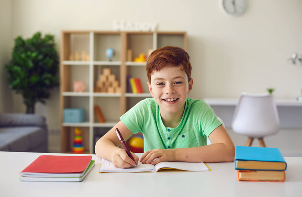 Online school. Smiling boy sitting at desk with notebooks and textbooks, doing homework and looking at camera over room interior background. - 写真・画像