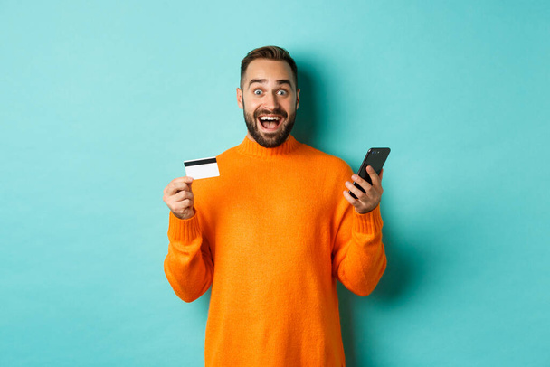 Online-Shopping. Überraschter Mann hält Handy und Kreditkarte in der Hand, bezahlt im Internet-Geschäft, steht vor hellblauem Hintergrund - Foto, Bild