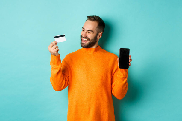Shopping online. Uomo soddisfatto utilizzando la carta di credito e mostrando schermo mobile, cercando contento, in piedi su sfondo azzurro - Foto, immagini