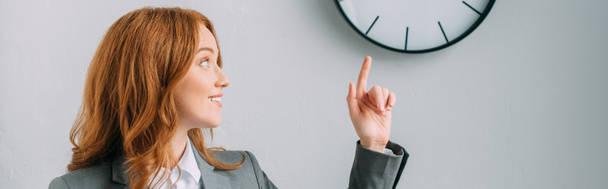 Positive redhead businesswoman pointing with finger and looking at wall clock on grey, banner - Fotoğraf, Görsel