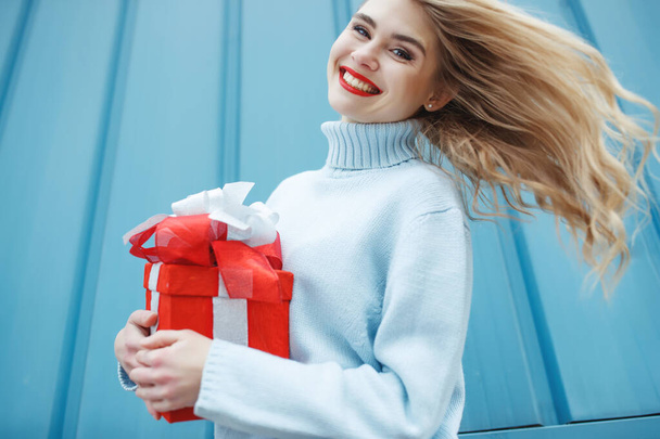 Щаслива блондинка тримає подарункову коробку і дивиться на камеру, а насолоджується синім фоном
. - Фото, зображення