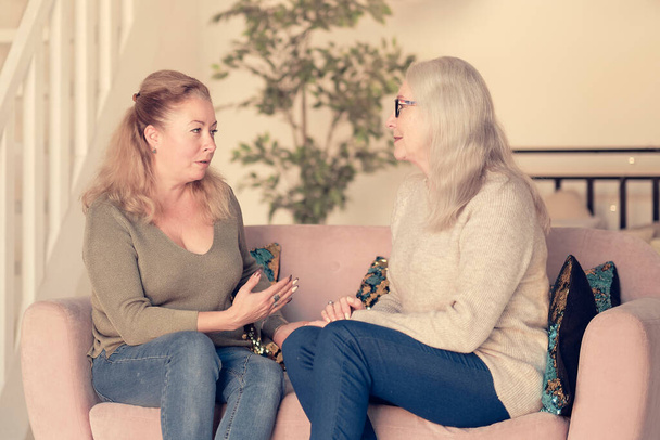 cute charming young female visiting her middle aged mother, sitting together on comfortable couch, having good conversation. Woman in her forties talking to daughter at home - Photo, Image