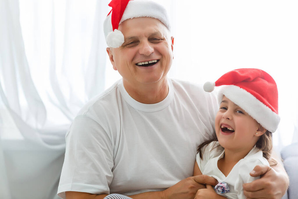 Retrato de cerca de una chica alegre abrazando al abuelo en la cabeza. abuelo y nieta en sombreros de Santa Claus - Foto, imagen