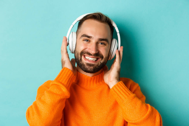 Κοντινό πλάνο του όμορφου σύγχρονου άνδρα που ακούει μουσική στα ακουστικά, στέκεται σε πορτοκαλί πουλόβερ πάνω από τυρκουάζ φόντο - Φωτογραφία, εικόνα