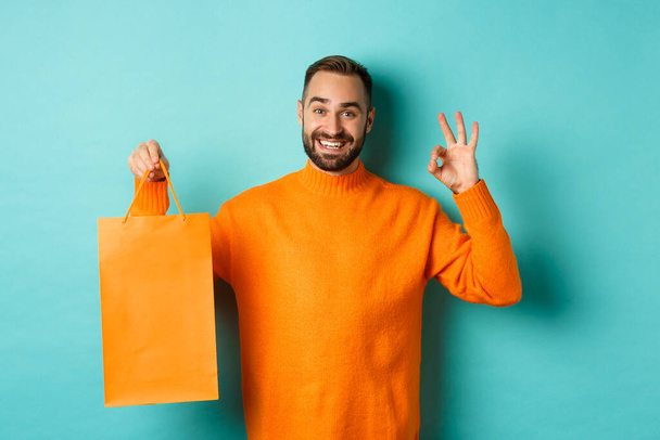 Satisfied male customer showing orange shopping bag and okay sign, recommending store, smiling pleased, standing over turquoise background - Photo, Image