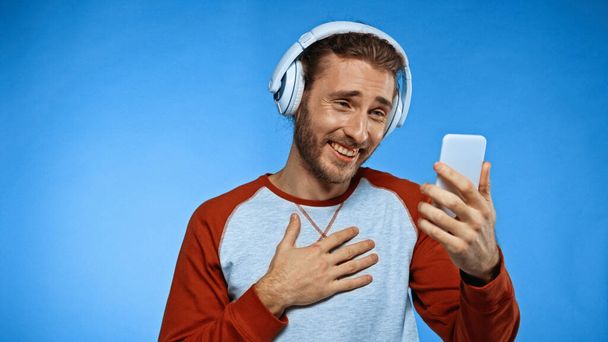 szczęśliwy człowiek w słuchawkach bezprzewodowych patrząc na telefon na niebiesko - Zdjęcie, obraz
