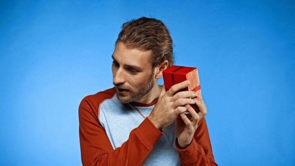 Neugieriger junger Mann schüttelt verpacktes rotes Geschenk auf blauem Grund - Foto, Bild