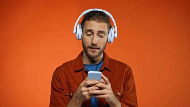 オレンジ色のスマートフォンを使ったワイヤレスヘッドフォンの髭の若い男  - 写真・画像