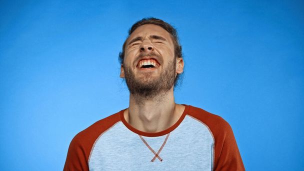 young bearded man with closed eyes laughing on blue  - Φωτογραφία, εικόνα