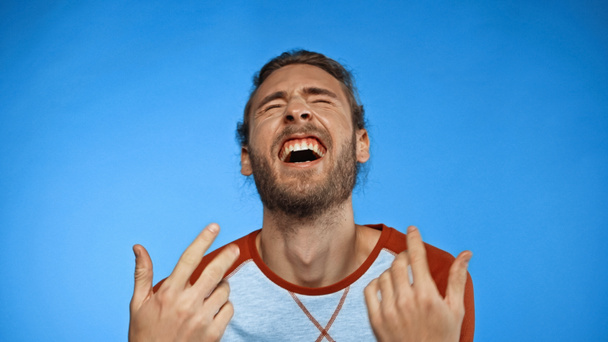 young bearded man with closed eyes gesturing while laughing on blue  - Foto, Bild