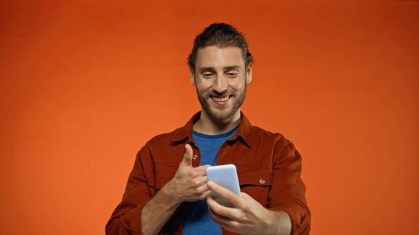 бородатый молодой человек показывает большой палец вверх, делая селфи на оранжевом  - Фото, изображение