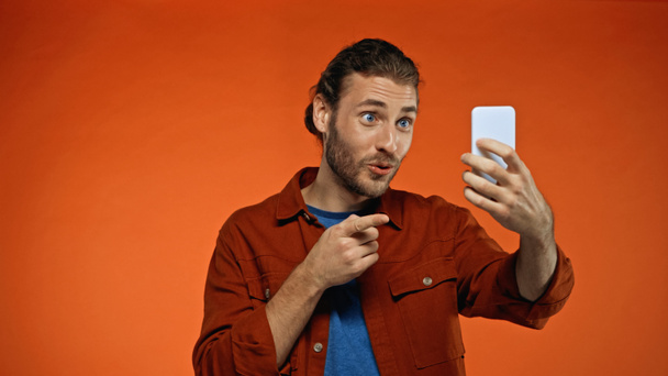 cheerful man pointing with finger while looking at smartphone and taking selfie on orange  - 写真・画像