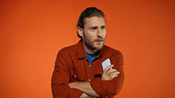bearded young man standing with crossed arms and holding phone on orange  - Photo, Image