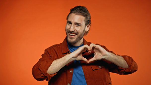 jeune homme positif souriant et montrant un signe cardiaque sur orange  - Photo, image