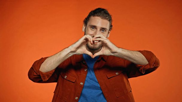 bearded young man smiling and showing heart sign on orange  - Photo, Image