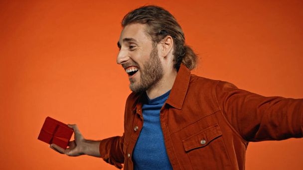 glücklicher junger Mann lächelt, während er ein Geschenk in Orange hält  - Foto, Bild