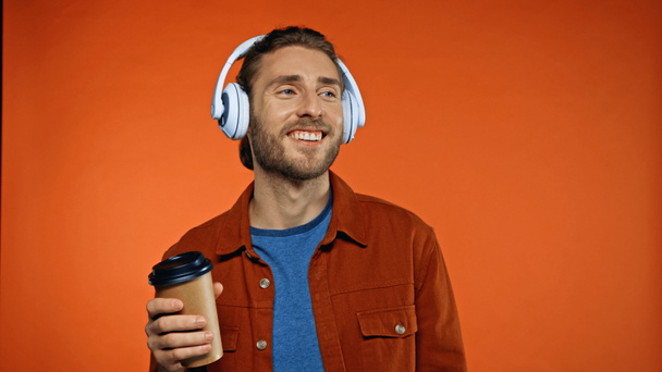 uomo allegro in cuffie wireless che tengono tazza di carta e ascoltare musica su arancione - Foto, immagini
