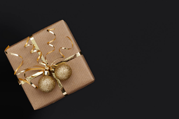 Christmas gift box or present decorated golden ribbon and two balls on black background. Flat lay - Φωτογραφία, εικόνα