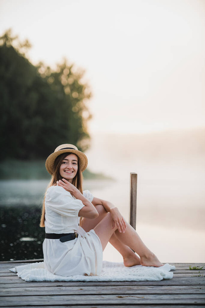 Attractive woman wearing white dress and hat sitting near lake. Warm and cozy summer morning atmosphere. - Foto, immagini