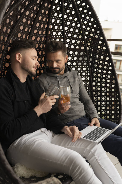 Cheers. Young handsome gay couple relaxing on the swing on the balcony with beer and browsing social media on laptop - Zdjęcie, obraz