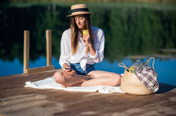 Beautiful young woman in hat has picnic near the lake in summer forest. She eats an apple and looking at smart phone. - 写真・画像