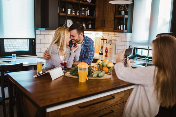 Девушка и ее прекрасные родители режут овощи и улыбаются во время приготовления пищи на кухне дома. Девушка фотографирует свою семью на смартфоне. - Фото, изображение