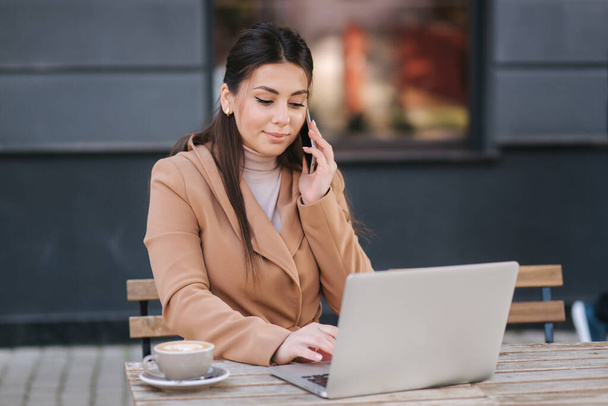 Young business woman sitting on terrace by cafe with laptop. Woman dring coffee and working outdoors. Female use phone and notebook - Foto, Imagem