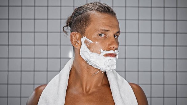 Мужчина без рубашки с белой пеной для бритья на лице смотрит в камеру в ванной комнате - Фото, изображение