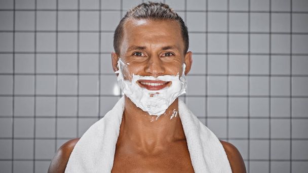 hombre positivo con espuma de afeitar en la cara mirando a la cámara en el baño - Foto, Imagen