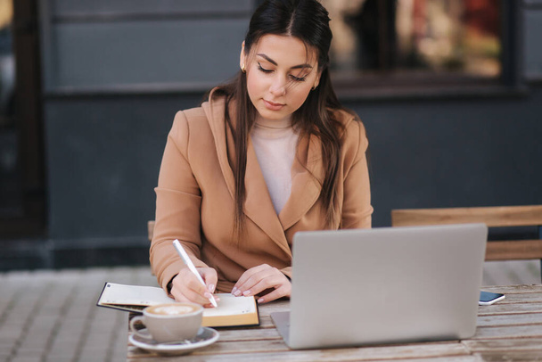 Atractiva joven empresaria escribiendo algo en un cuaderno. Beber café femenino y trabajar al aire libre con computadora portátil - Foto, imagen