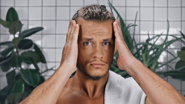 Мужчина без рубашки регулирует волосы возле зеленых растений на размытом фоне в ванной комнате  - Фото, изображение