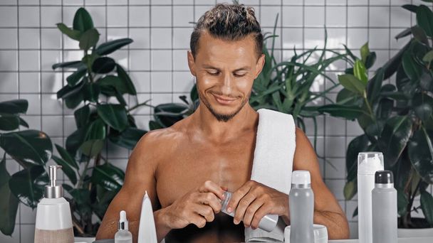 homem feliz segurando garrafa com espuma de barbear perto de plantas verdes no fundo borrado no banheiro  - Foto, Imagem
