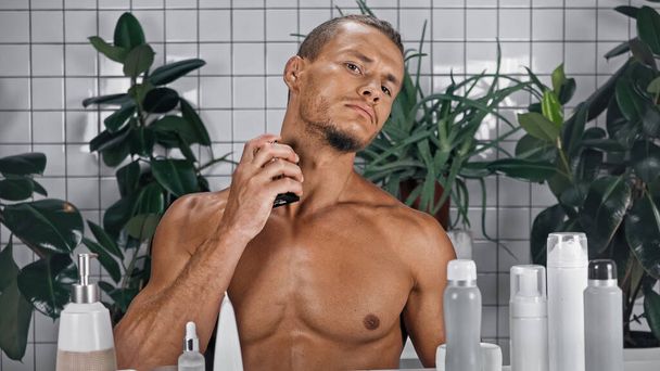 félmeztelen férfi permetezés parfüm közelében zöld növények elmosódott háttér fürdőszobában  - Fotó, kép