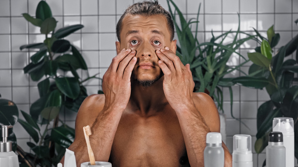 молодий без сорочки чоловік торкається обличчя біля зелених рослин на розмитому фоні у ванній
  - Фото, зображення